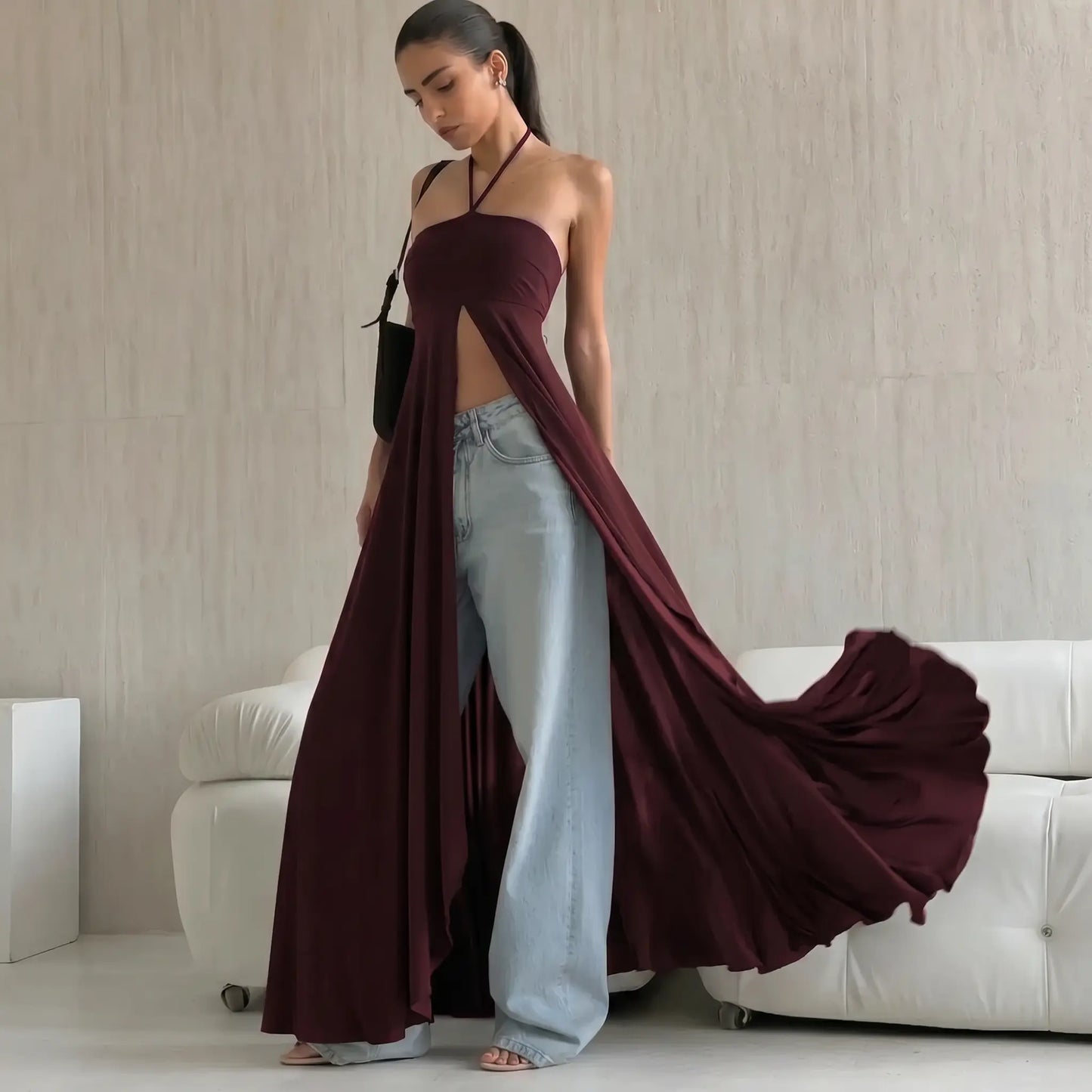 A woman models a deep chocolate brown, flowing maxi dress with a halter neckline and dramatic high front slit, styled stylishly over light-wash wide-leg jeans and sandals, set against a chic interior with a white sofa.