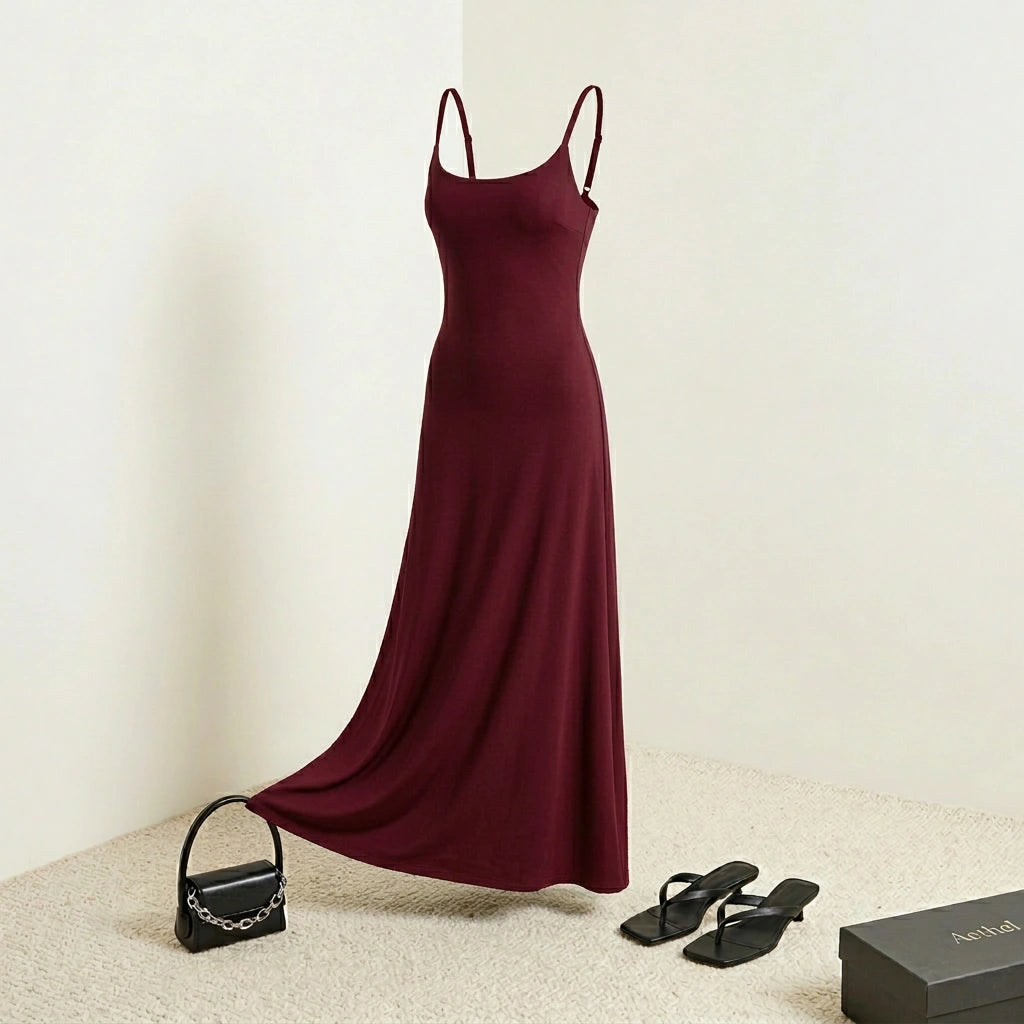 Aethel sleek maxi dress in deep burgundy with adjustable spaghetti straps, styled with a handbag and sandals in a minimalist room.