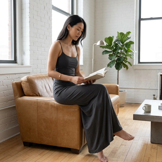 Aethel charcoal grey sleek maxi dress with form-fitting bodice and flowing skirt, worn by a woman reading in a bright studio.