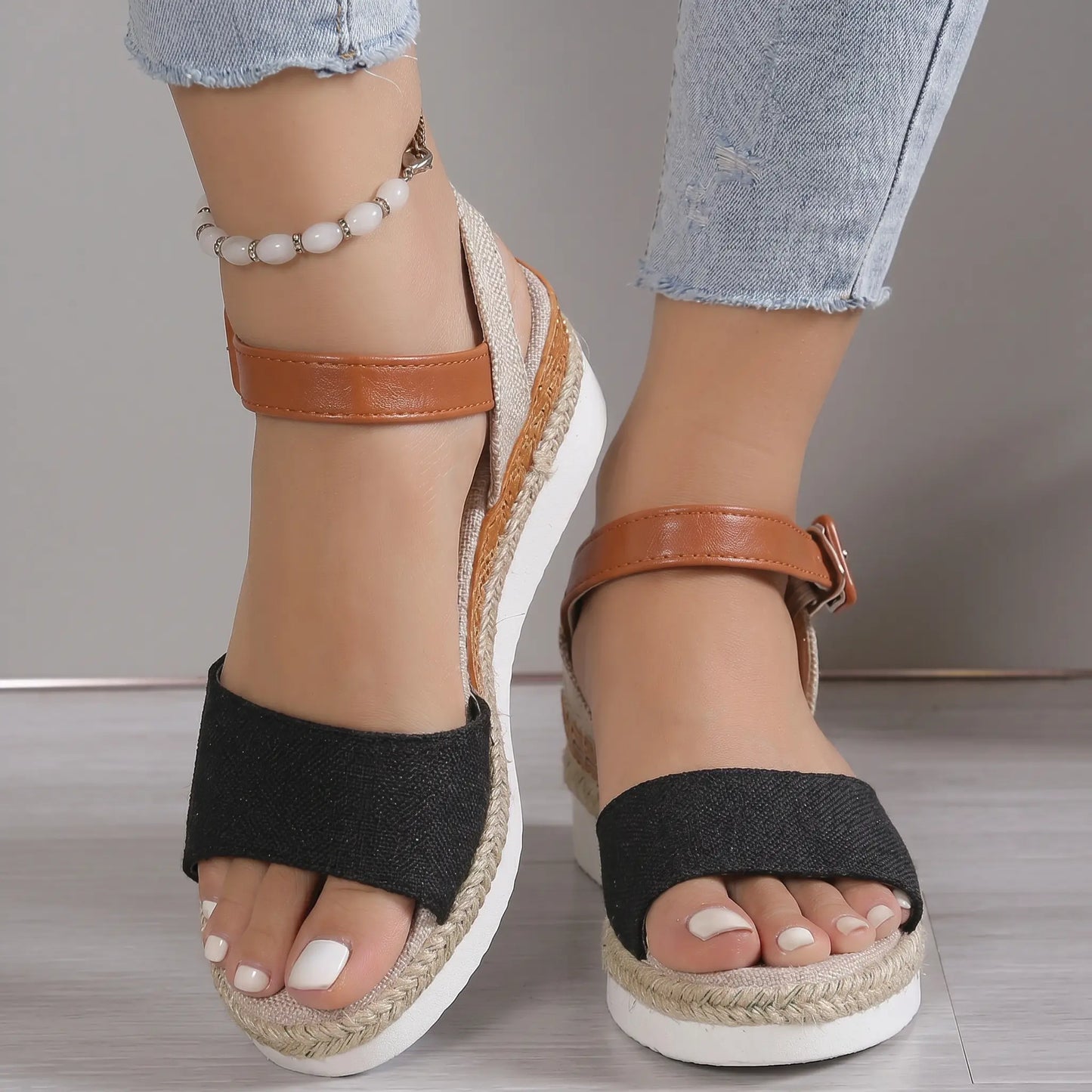 Women's black espadrille wedge sandals with tan faux leather ankle straps shown on a model's feet with light blue jeans. The shoes feature a peep-toe and jute-wrapped wedge heel.