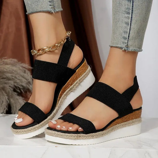 Black espadrille wedge sandals with peep-toe, textured mesh upper, elasticated straps, and a gold-tone ankle chain, worn by a model.
