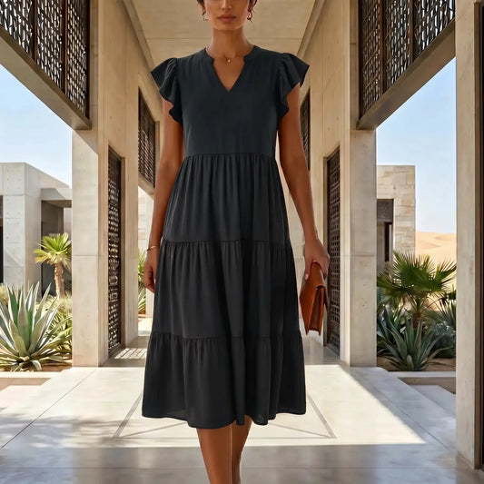 Model in black Torvun ruffled tiered midi dress with V-neck and flutter sleeves, walking through an elegant architectural hallway.