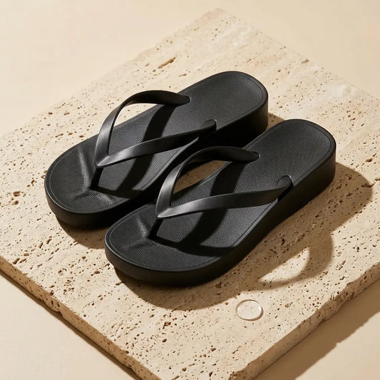 A pair of black waterproof PVC platform wedge flip-flops with textured footbeds and sleek Y-straps, displayed elegantly on a sunlit, textured stone slab.