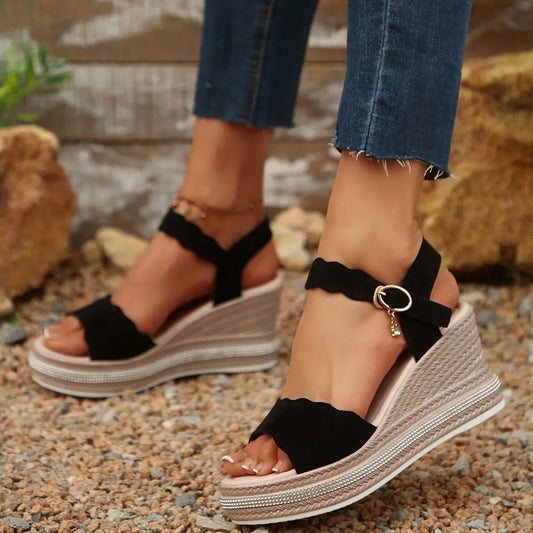 Black suede-like wedge sandals featuring scalloped straps and a rhinestone-embellished platform, worn on a woman's feet in an outdoor, gravelly setting with natural foliage.