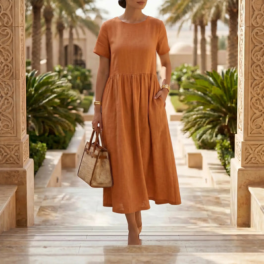 Elegant model wearing a Kivori burnt orange linen-blend midi dress with a relaxed A-line silhouette and short sleeves, walking outdoors with a straw tote bag.