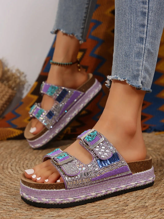 Woman's feet wearing embellished platform slide sandals with purple straps, rhinestones, beads, and blue resin stones, paired with denim jeans.