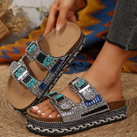 Embellished platform slide sandals with double buckle straps, featuring intricate rhinestones, beads, and vibrant turquoise resin stones, shown on a woman's feet in a boho-chic setting.