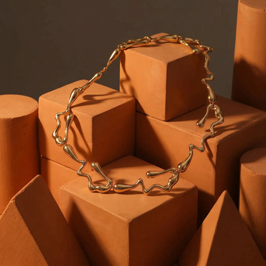 High-polish gold liquid metal choker necklace featuring wavy, irregular links, displayed on terracotta geometric blocks in a studio setting.