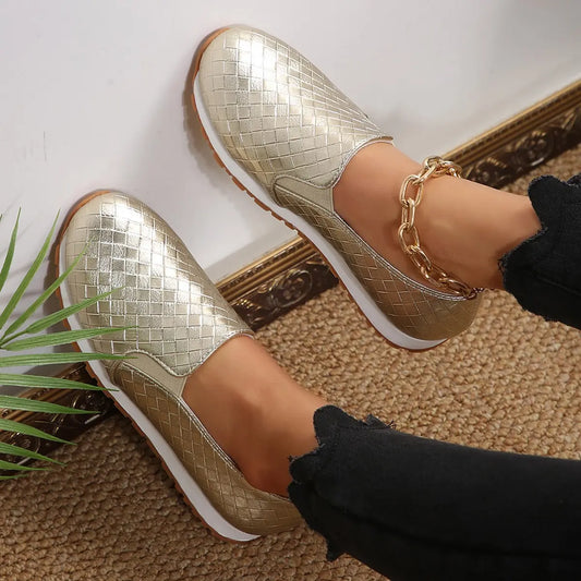 Model's feet wearing metallic gold woven slip-on loafers with a gold chain anklet and black ripped jeans.