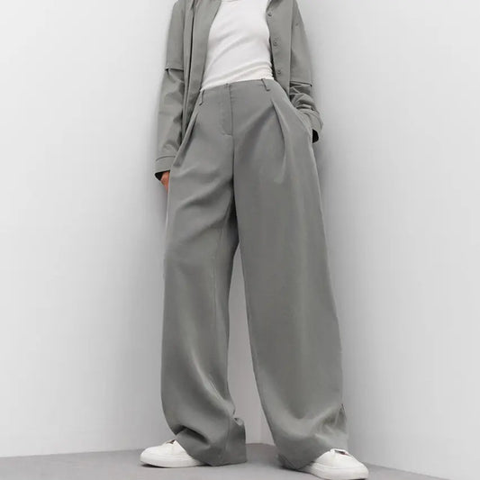 Sophisticated high-waisted wide-leg trousers in a muted grey, featuring sharp front pleats and a fluid drape, paired with a white top and sneakers.