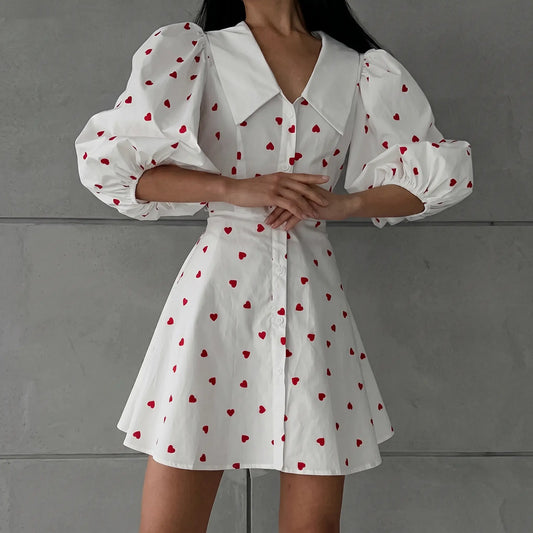 Woman wearing a white cotton linen blend A-line midi dress with a red heart print, featuring dramatic puff sleeves and a pointed collar, shown from the front.