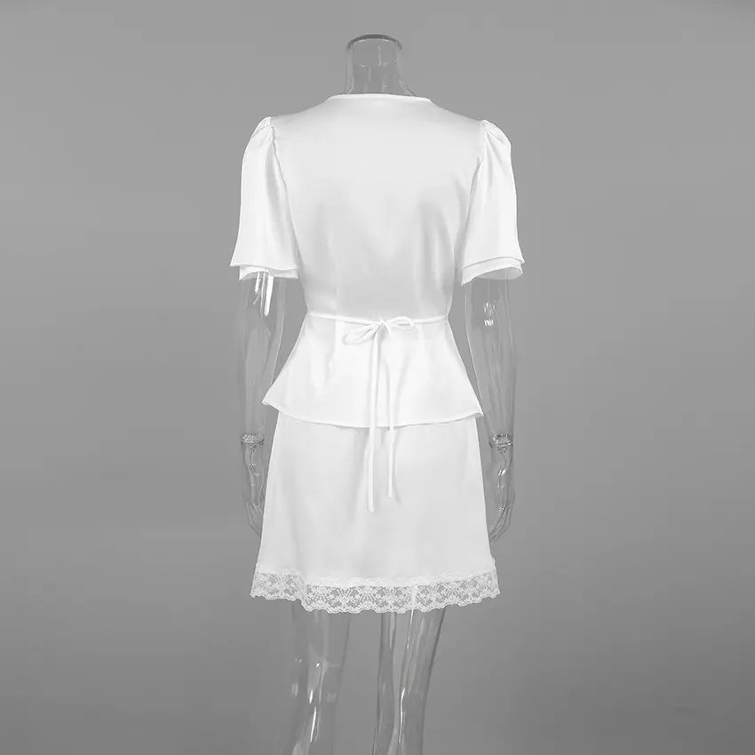 Back view of an ivory satin two-piece set featuring a short-sleeved blouse with puffed sleeves and a lace-trimmed mini skirt. The blouse has a drawstring tie at the waist and a delicate lace hem on the skirt.