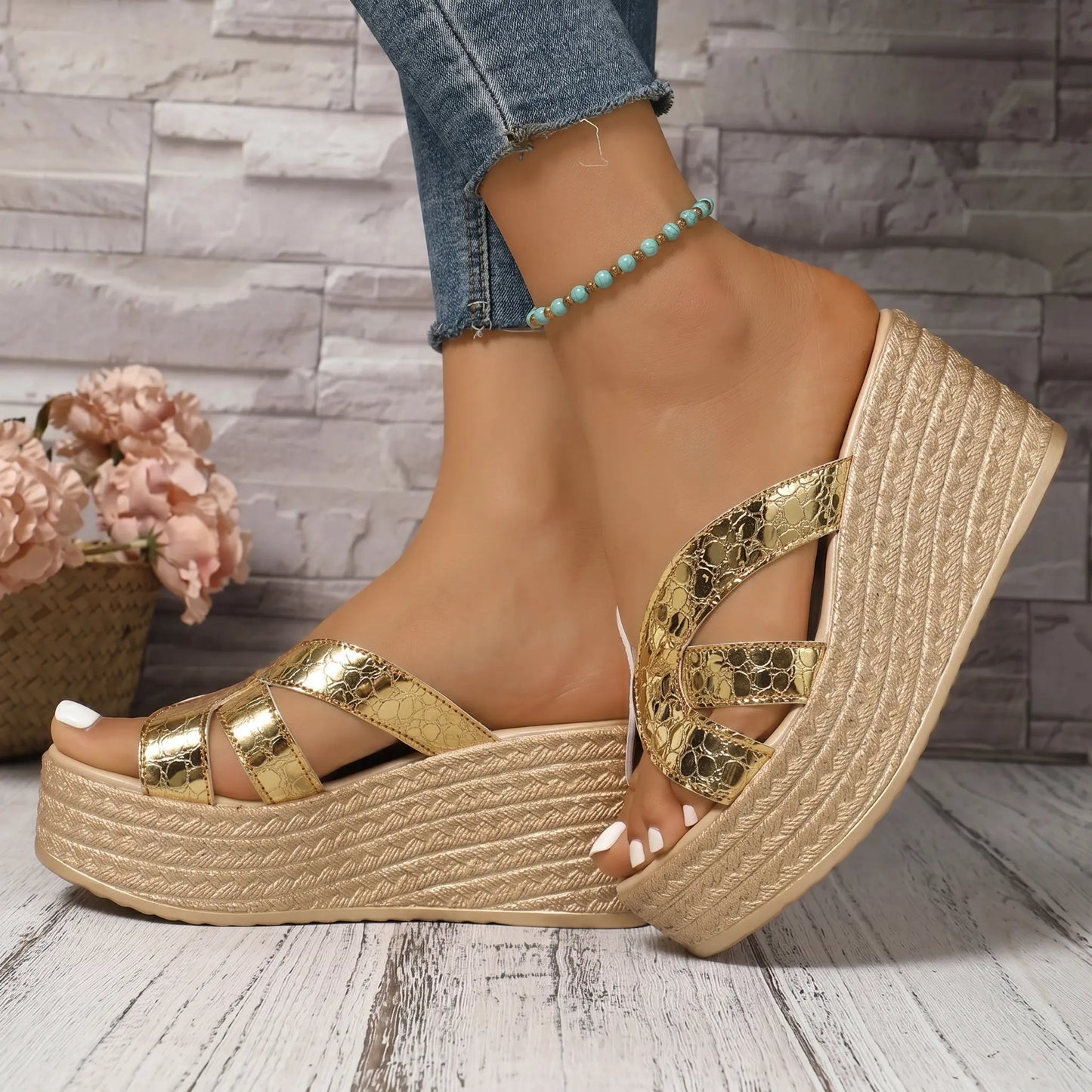 Kaelen gold metallic wedge espadrille sandals featuring criss-cross straps and a thick platform sole, shown from a three-quarter angle on a wooden floor with a brick wall backdrop.