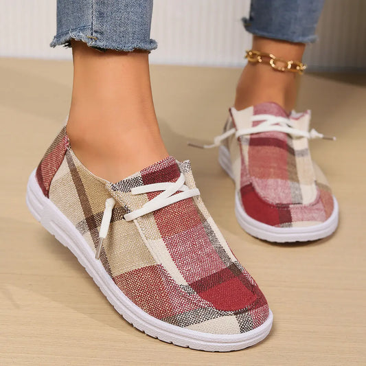 Close-up lifestyle shot of Kaelen plaid canvas loafers worn with blue jeans, showcasing the red, beige, and brown plaid pattern and white laces against a warm wooden floor.