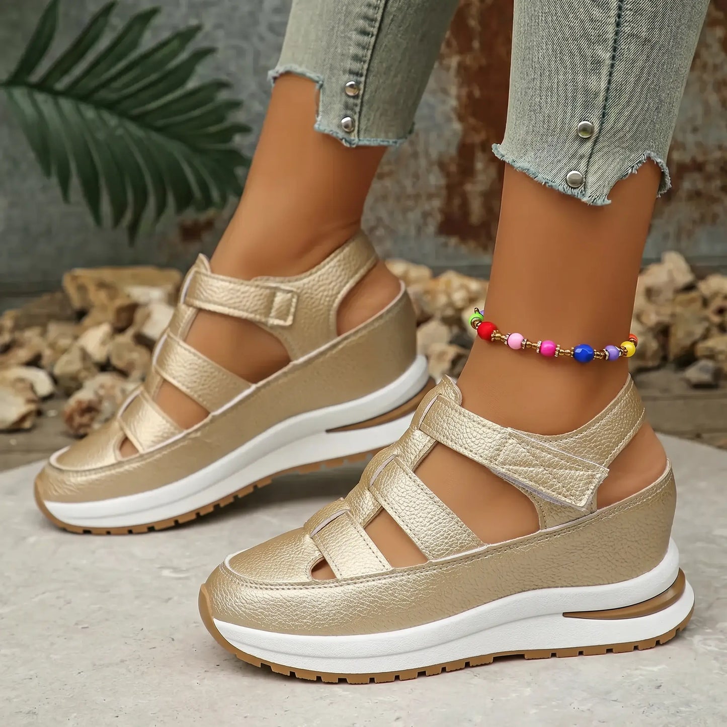 Kaelum women's subtle gold metallic platform wedge sandals with cut-out straps are shown on a model's feet with cropped jeans and a colorful anklet, on a textured backdrop.