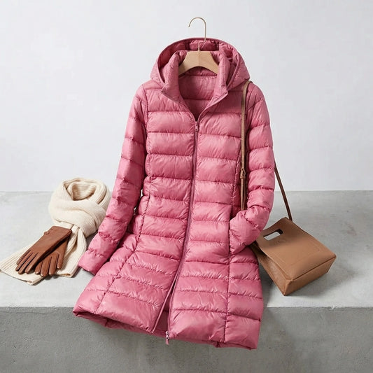 Kovera long puffer coat in soft rose pink with removable hood, styled with a scarf, gloves, and handbag on a concrete surface.