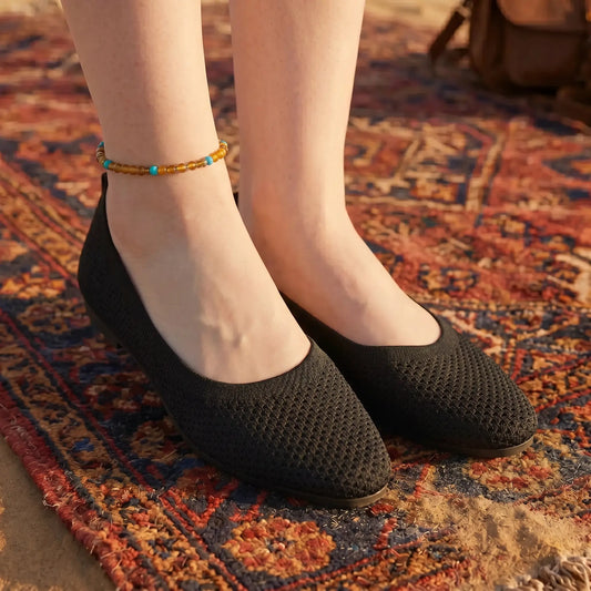A person wears black Kyneth breathable knit pointed flats with an anklet, standing on a vibrant bohemian rug outdoors.