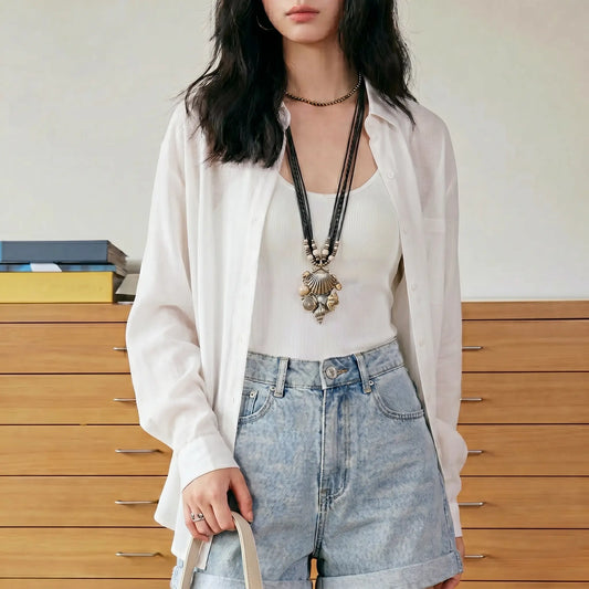 Female model wearing a Kynzor Cloud White Tencel button-down blouse open over a white tank top, paired with denim shorts, showcasing the relaxed fit and semi-sheer fabric.