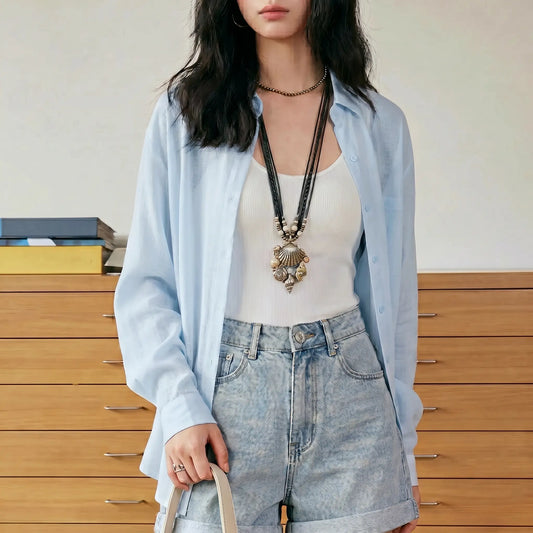 Female model wearing the Kynzor Breeze Light Blue Lightweight Tencel Button-Down Blouse, styled open over a white tank top with light wash denim shorts in a bright indoor setting.