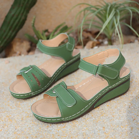 Green Lyraia comfort wedge sandals with peep-toe front and adjustable hook-and-loop straps shown in a natural outdoor setting with sand and plants.