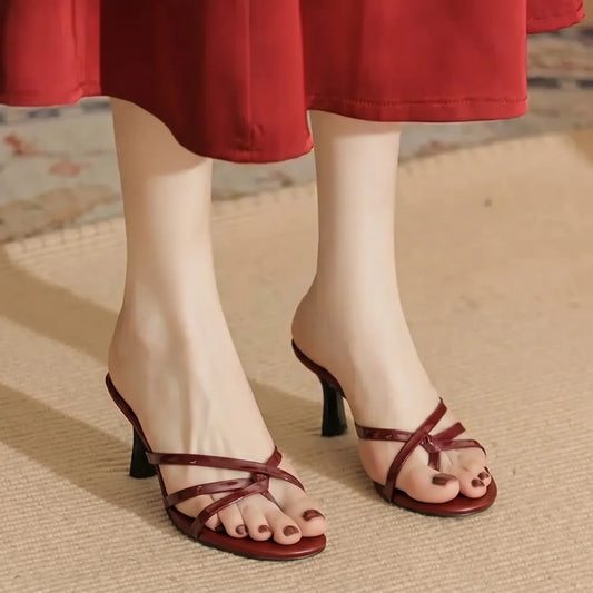 Elegant oxblood red strappy stiletto mules worn by a model, showcasing delicate criss-cross straps, an open almond toe, and a slender heel, styled with a flowing red dress.