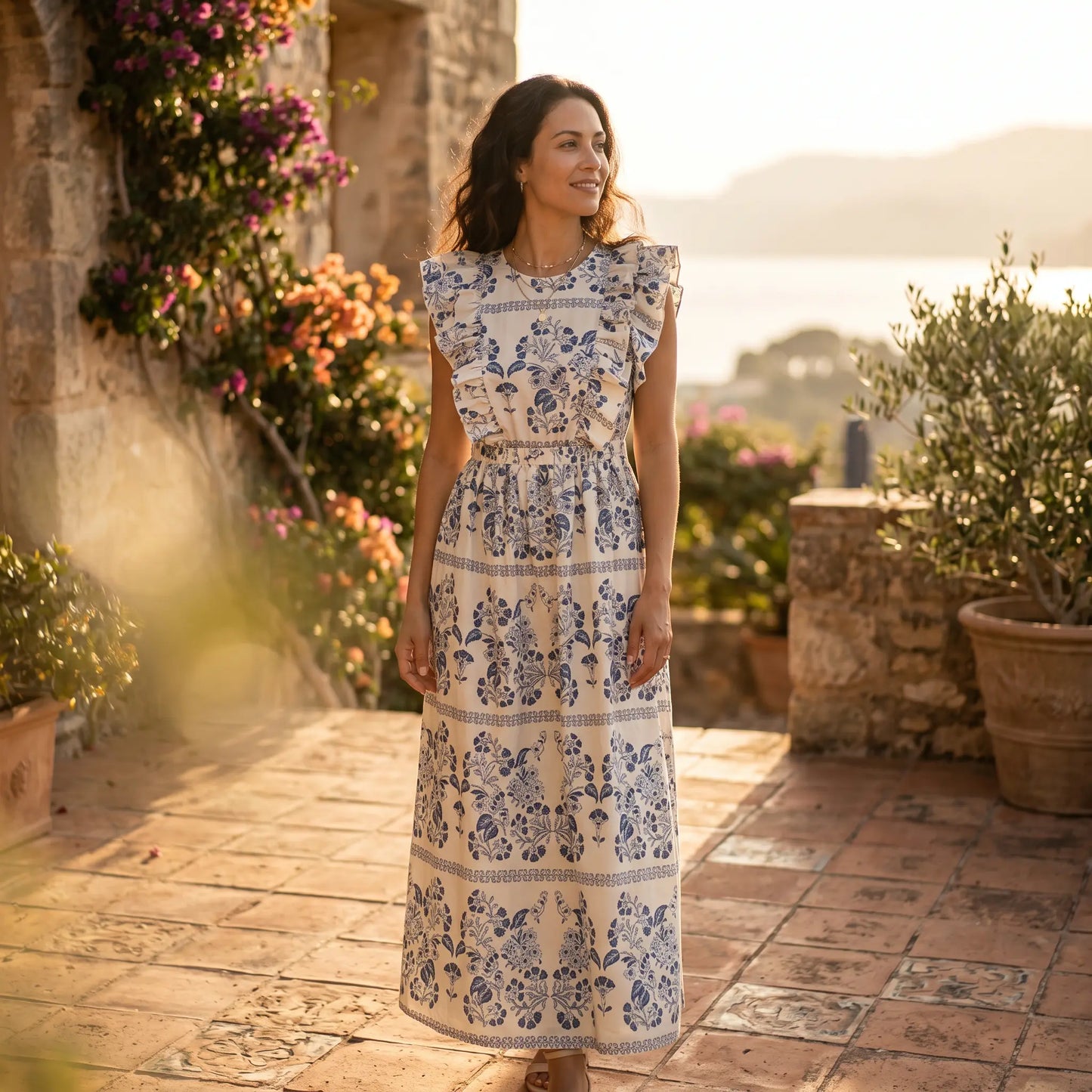 A woman models a white floral maxi dress with blue botanical print, ruffled cap sleeves, and an elasticated waist, standing outdoors in a sunny, picturesque setting with stone walls and flowering plants.