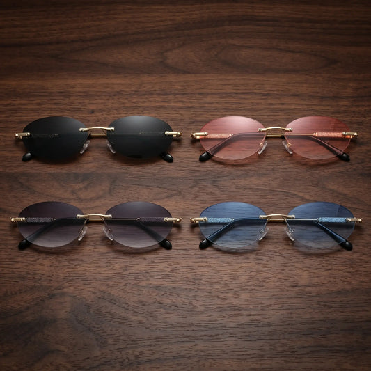 Four pairs of Nymira oval rimless sunglasses with gold-tone metal frames and gradient lenses in black, pink, grey, and blue tints, laid out on a wooden surface.