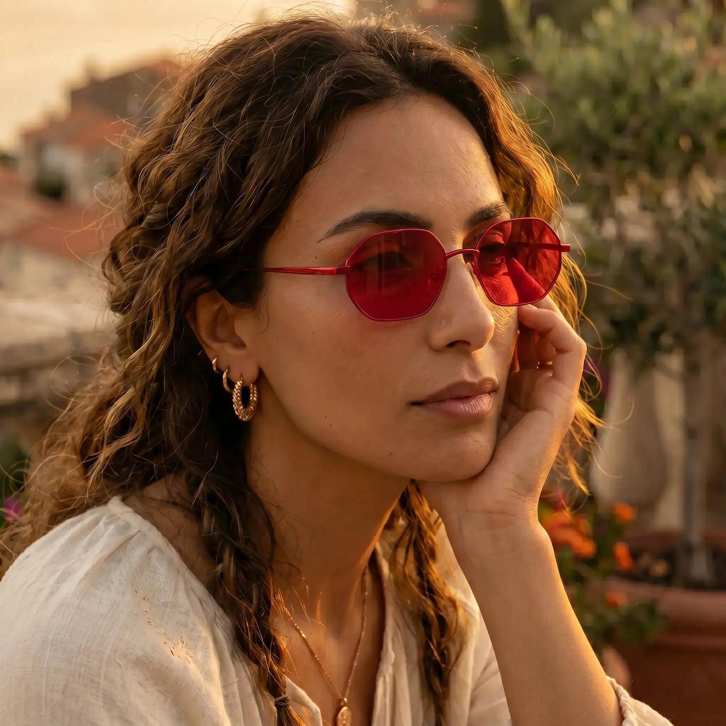 A woman with curly brown hair wears vibrant crimson octagonal sunglasses with thin gold-tone metal frames, set against a sun-drenched outdoor backdrop with blurred buildings and foliage.