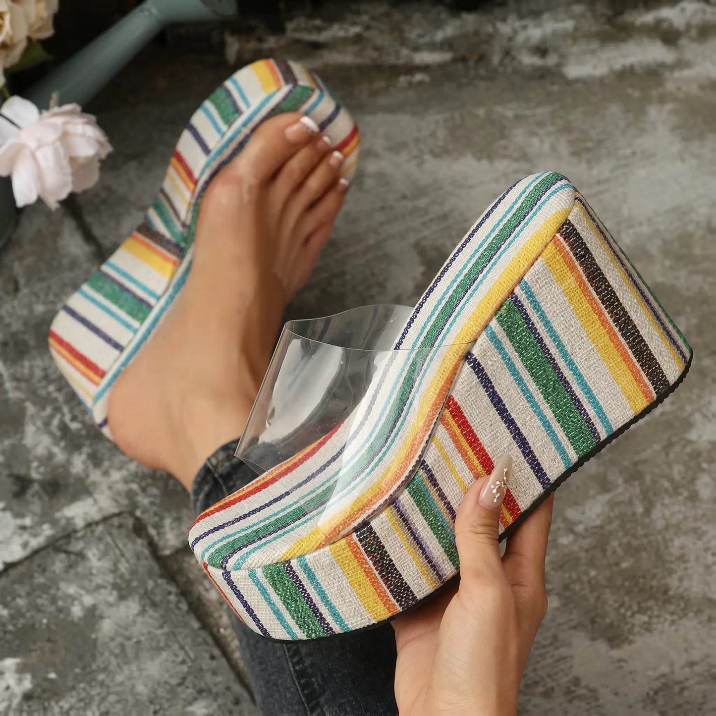 Close-up of a woman's feet wearing vibrant, multi-colored striped platform wedge sandals with a clear PVC strap, set against a textured outdoor ground.