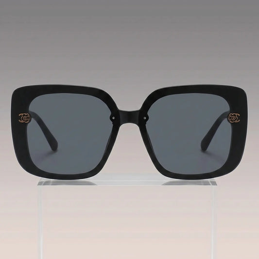Vesryn oversized square sunglasses with thick black acetate frames and dark gradient grey lenses, featuring gold floral-inspired metal logo accents on the temples.