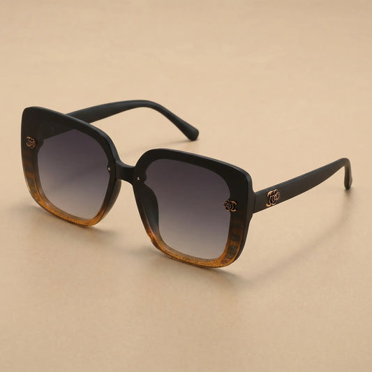 Oversized square sunglasses with a matte black frame, featuring a gradient amber-brown front and dark gradient grey lenses. Embellished with a gold floral-inspired logo on the temples and subtle textured detail on the frame corners.