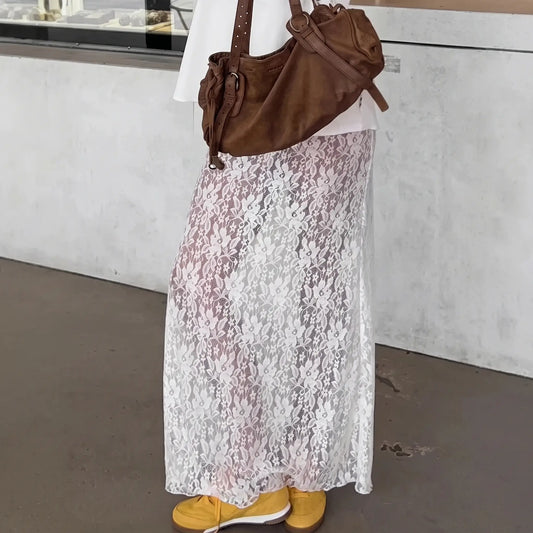 A woman wearing a floor-length white floral lace A-line maxi skirt, styled with a white top, brown shoulder bag, and yellow sneakers against a textured concrete wall.