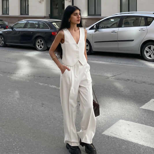 A woman models a crisp white V-neck sleeveless vest and fluid wide-leg trousers ensemble, styled in an urban outdoor setting with cars and buildings visible.