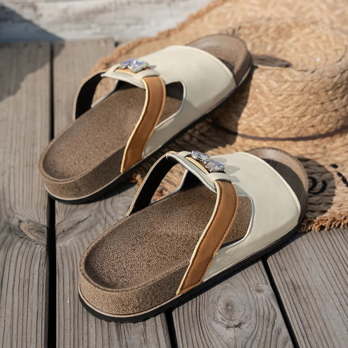 Xylia embellished flat slide sandals in cream and tan, featuring a clear crystal butterfly accent and a cork footbed, styled on a straw hat and wooden deck.