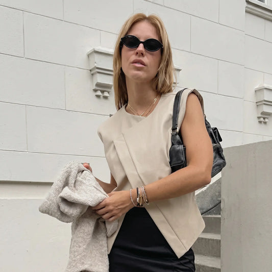 Woman wearing a sleeveless Xylohn asymmetric drape top in soft champagne beige, styled with a black skirt and handbag, against an urban textured wall background.
