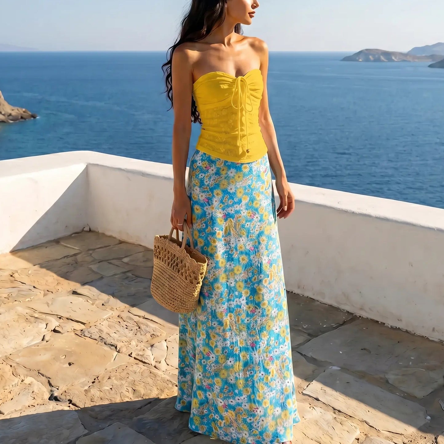 Female model wearing Zendri sunlight yellow ruched strapless top and cerulean blue floral maxi skirt, standing on a sunny patio overlooking the ocean.