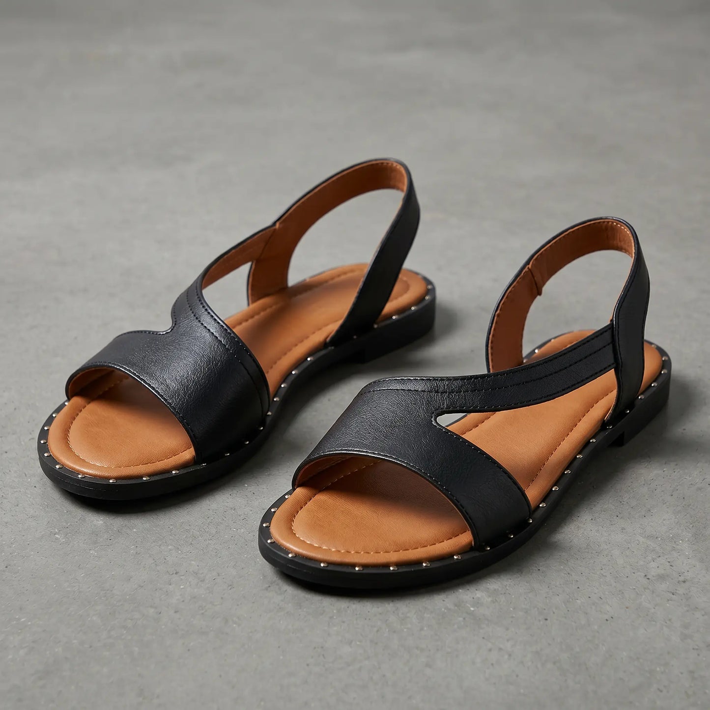 Black faux leather Zylos Roman flat sandals featuring a geometric strap design, delicate ankle strap, and metallic sole studs, shown from a three-quarter angle on a textured grey studio background.