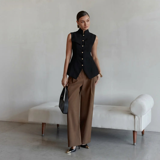 Woman wearing a tailored black sleeveless vest with a high mandarin collar and polished gold buttons, paired with taupe trousers and a black handbag.