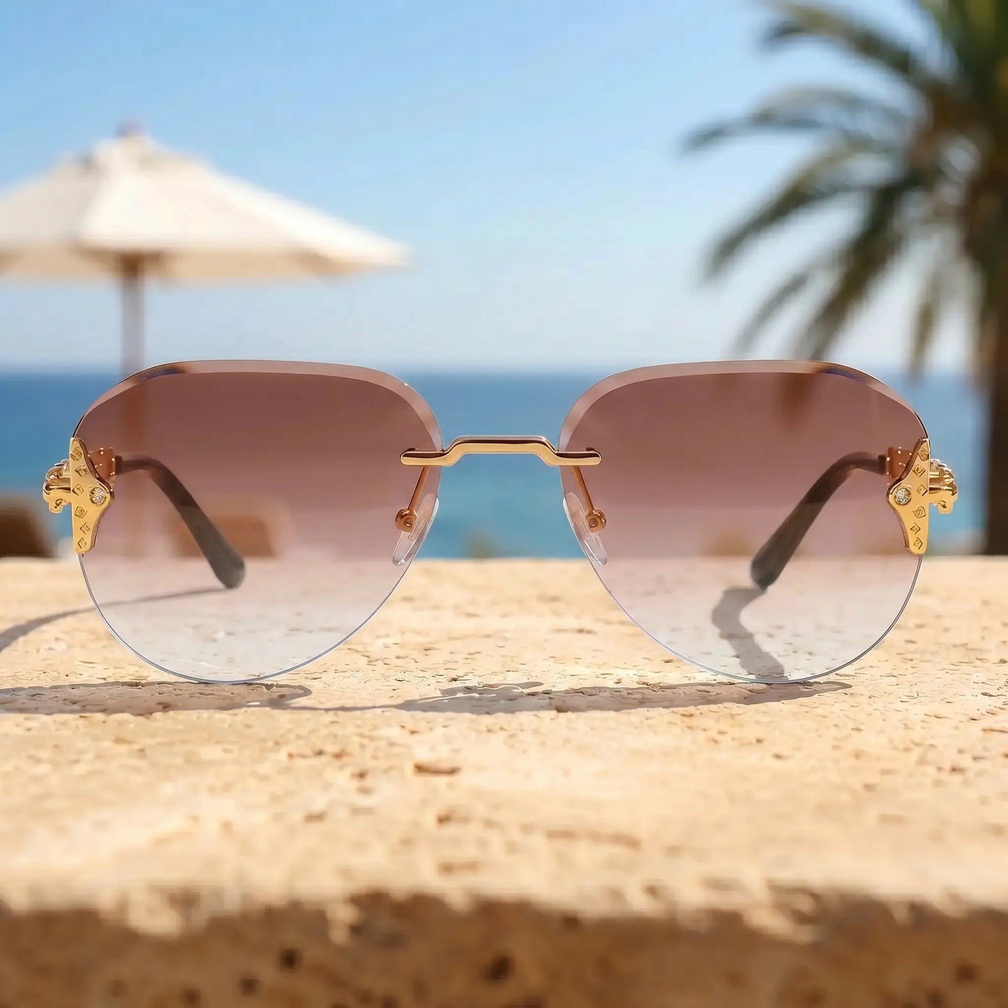 Gold-tone rimless aviator sunglasses with ornate chain-link temples and brown pink gradient lenses, styled on a textured surface with a beach and ocean backdrop.