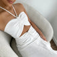 A woman models a white Aelyra halterneck maxi dress with a ruched bust, midriff cut-out, and drawstring waist, seated in a textured armchair for a sophisticated lifestyle shot. The breathable cotton-linen blend fabric offers effortless elegance.