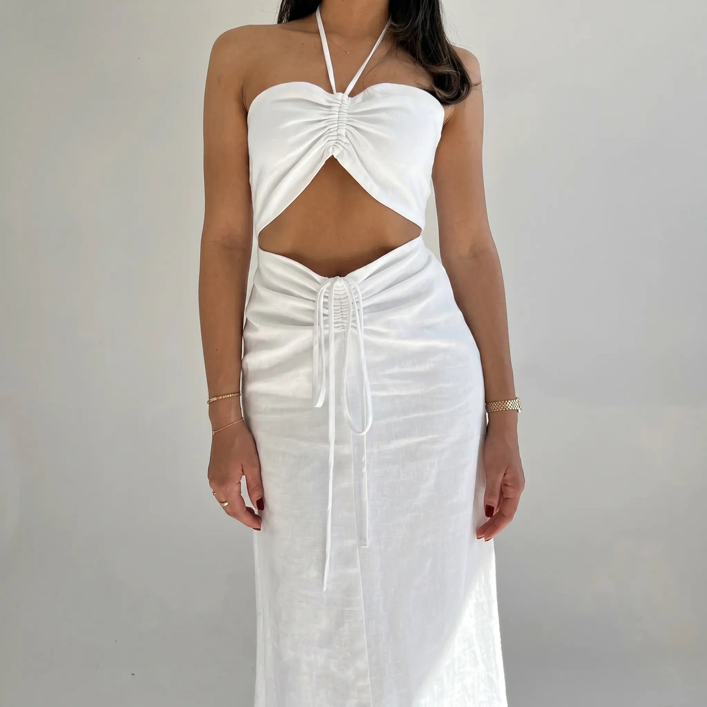 Aelyra white cotton-linen blend halterneck maxi dress with ruched bust detailing, a midriff cut-out, and adjustable drawstring waist, shown front view on a model against a light grey studio background.