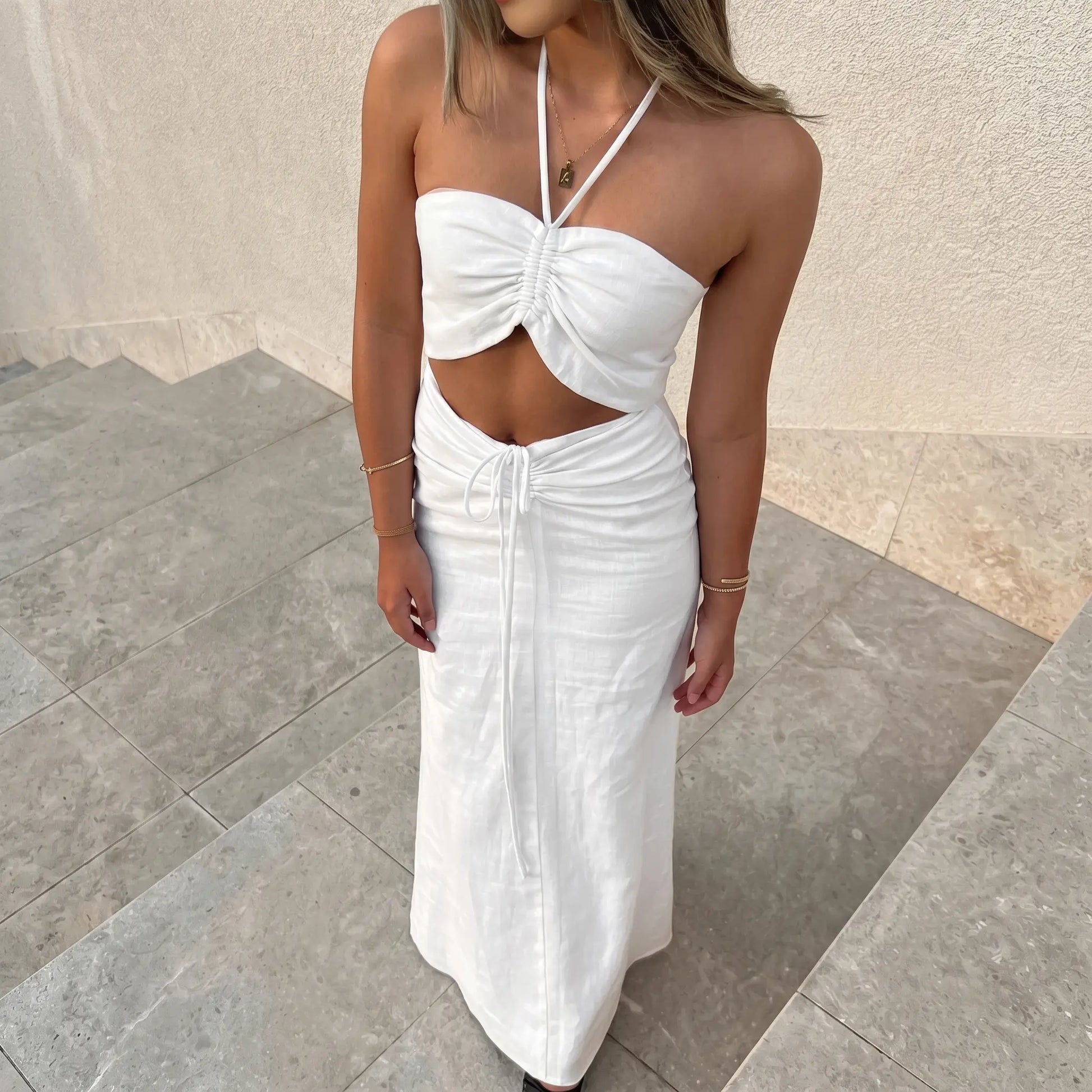 Aelyra white ruched halterneck maxi dress with midriff cut-out and drawstring waist, worn by a model on outdoor stairs.