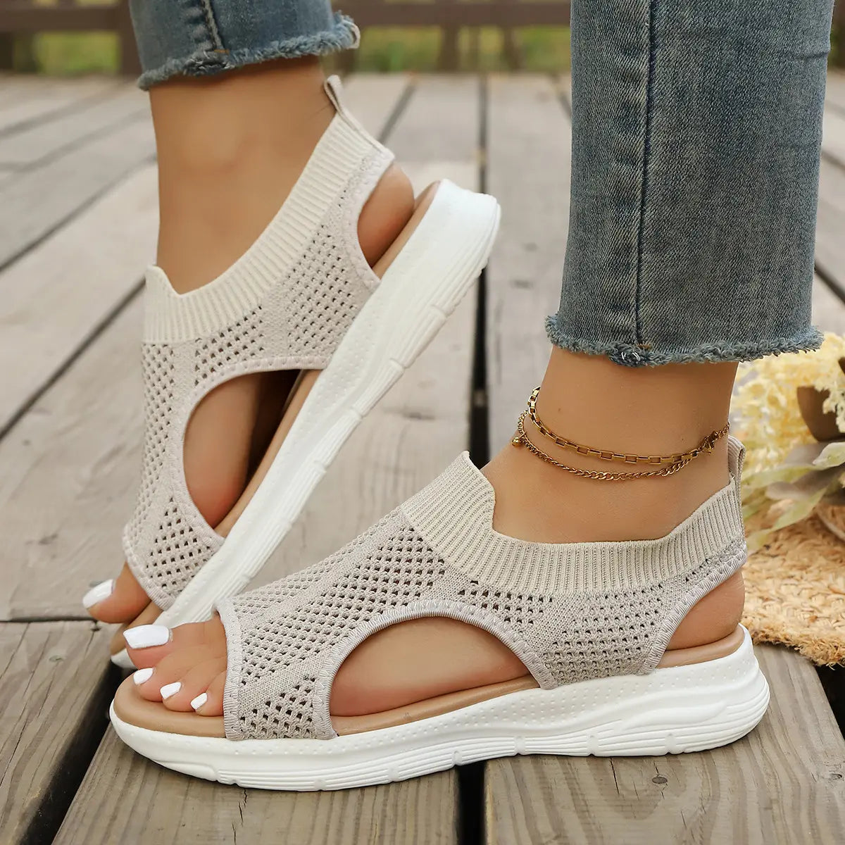 Woman wearing soft beige breathable knit mesh slip-on sandals with an open toe and elasticated heel strap, styled with denim jeans and an ankle bracelet on a wooden deck.