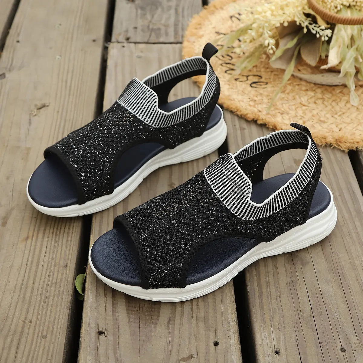Pair of black Aeriva women's breathable knit sandals with white elasticated heel straps and layered foam soles, displayed on a wooden deck with a straw hat.