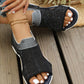 Black breathable knit slip-on sandals with an elasticated heel strap, worn by a woman with white-painted toenails and a gold ankle bracelet, styled on a wooden deck with a straw hat and greenery in the background.