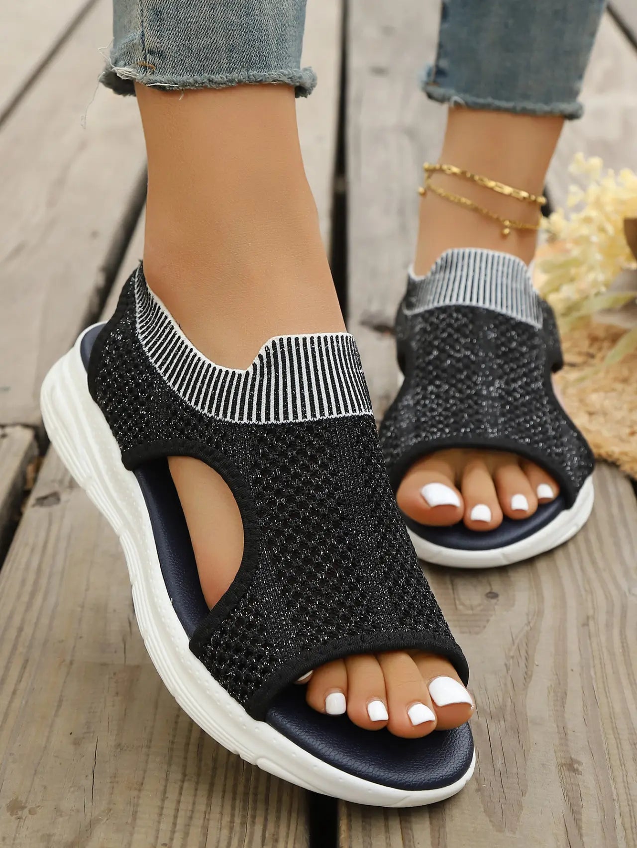 Women's black breathable knit mesh sports sandals with an open toe, layered white sole, and elasticated heel strap, shown on feet styled with distressed denim and an anklet.