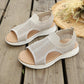 A pair of Aeriva women's breathable knit sandals in soft beige are displayed on a wooden deck, styled with a straw hat and dried flowers. These slip-on flat sandals feature an intricate mesh upper, cushioned footbed, and elasticated heel strap for comfort and a bohemian-chic look. Perfect for summer wear.