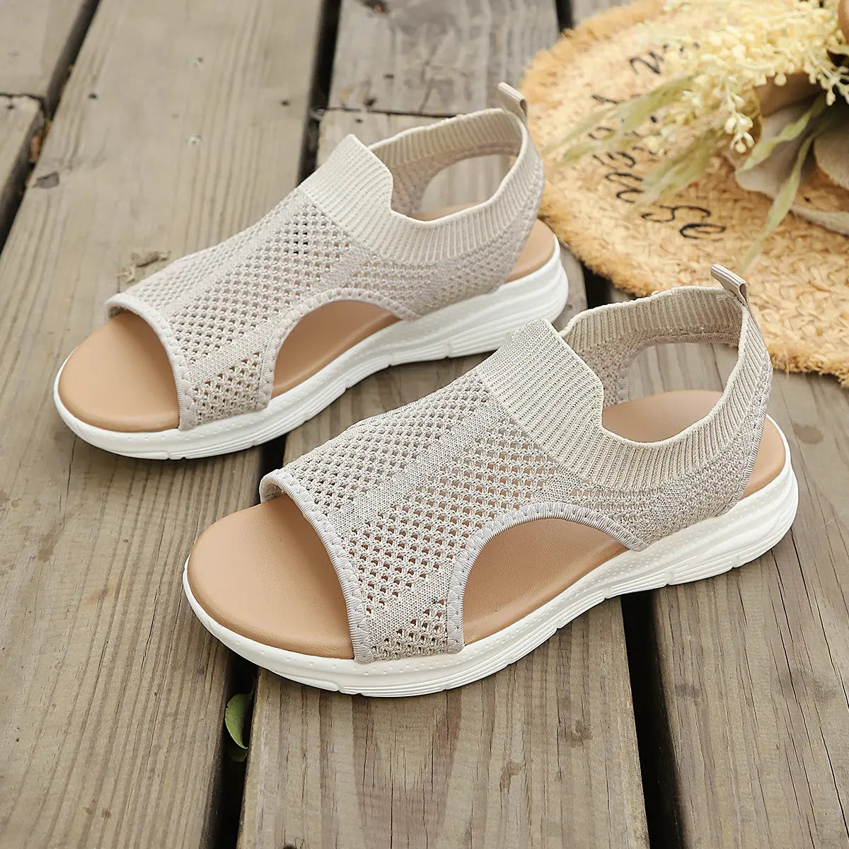 A pair of Aeriva women's breathable knit sandals in soft beige are displayed on a wooden deck, styled with a straw hat and dried flowers. These slip-on flat sandals feature an intricate mesh upper, cushioned footbed, and elasticated heel strap for comfort and a bohemian-chic look. Perfect for summer wear.