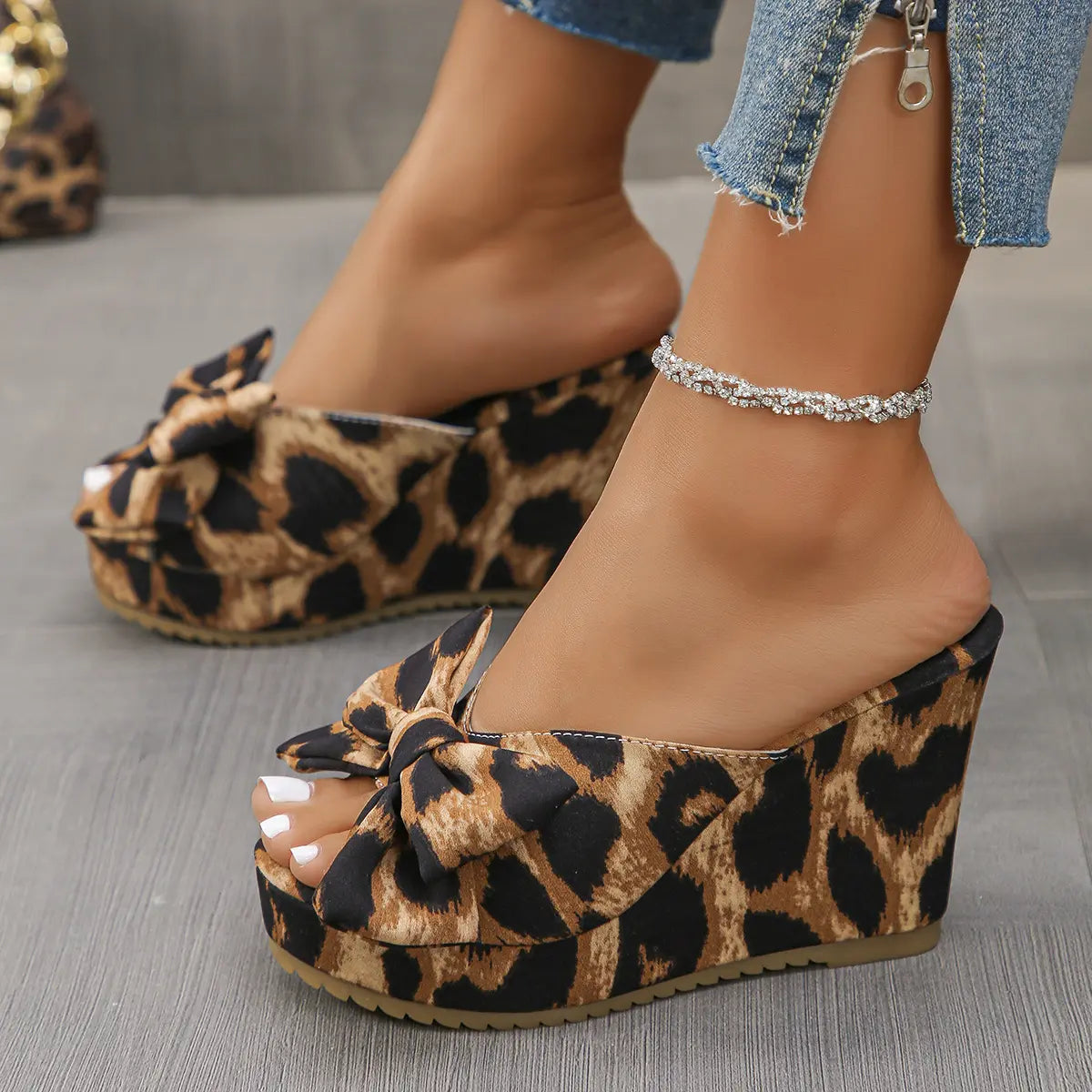 Aerona leopard print wedge sandals with an oversized bow and peep-toe, worn by a model with jeans and an anklet, showcasing the side and front profile on a grey floor.