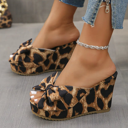Aerona leopard print wedge sandals with an oversized bow and peep-toe, worn by a model with jeans and an anklet, showcasing the side and front profile on a grey floor.